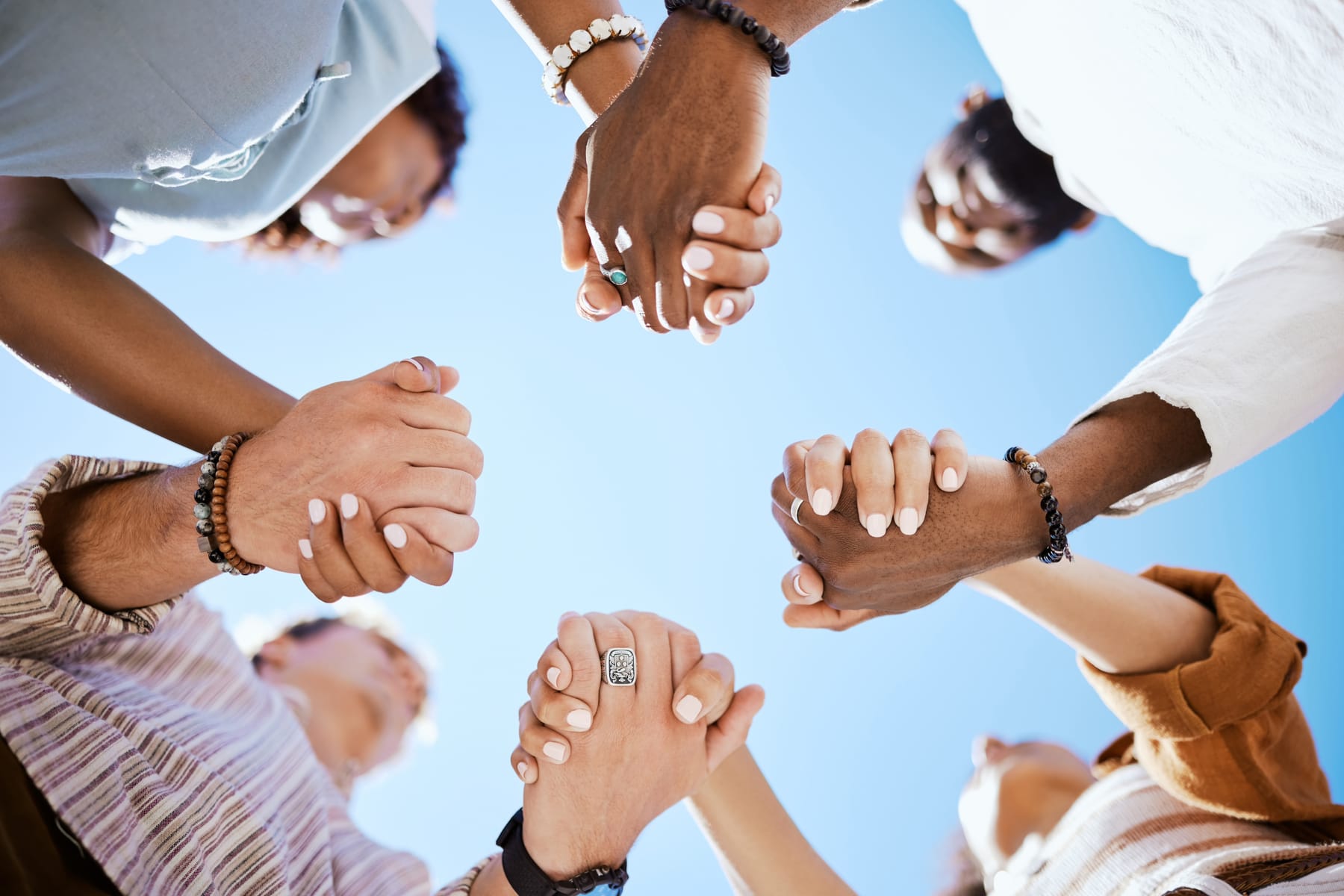 A circle of people holding hands, seen from below against a bright blue sky