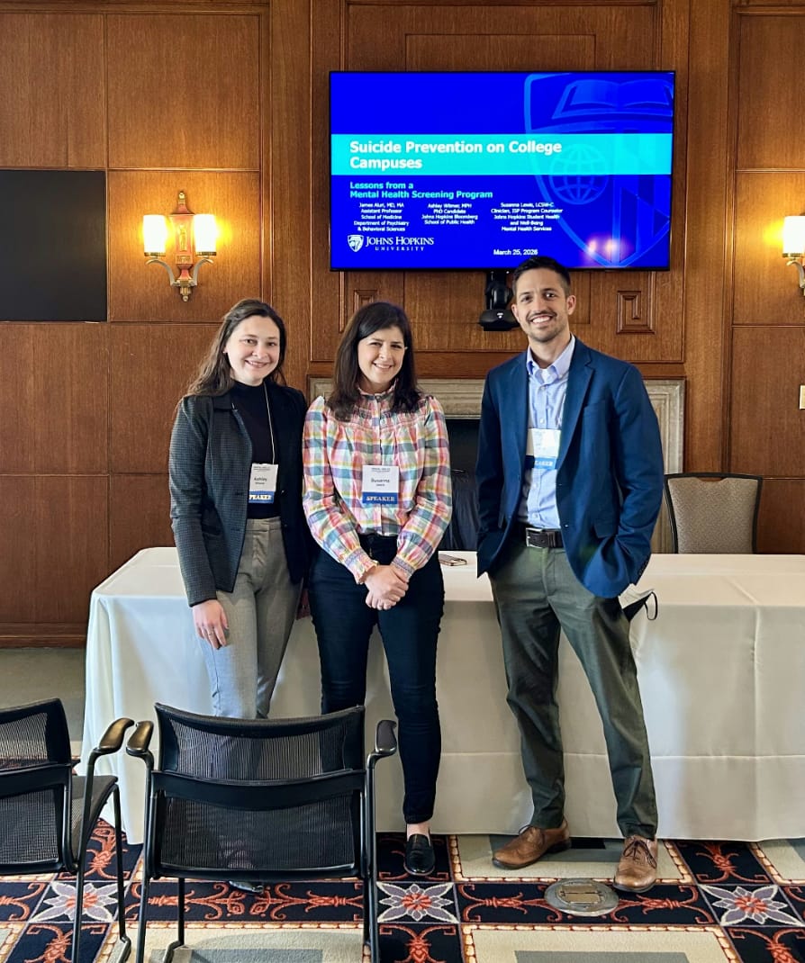 Three presenters stand in front of a screen showing their talk, “Suicide Prevention on College Campuses: Lessons from a Mental Health Screening Program.”